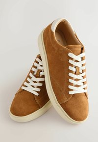 Paire de baskets en daim marron avec lacets blancs et semelles en caoutchouc crème sur un fond clair uni.