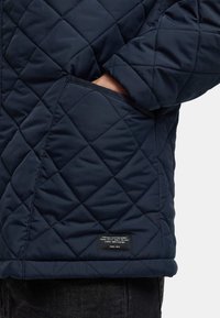 Marineblau gesteppte Jacke mit einer Patentedasche, strukturiertem Stoff und einem Etikett nahe dem Saum mit Markendetails.