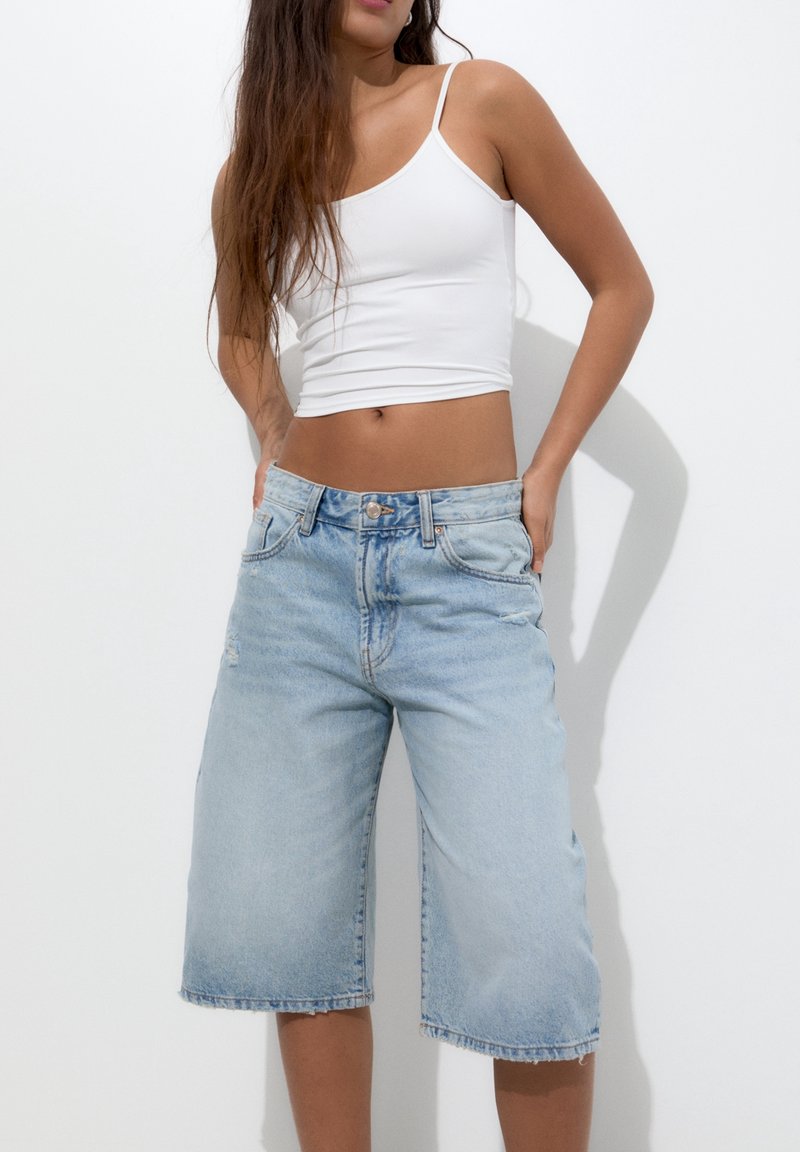 Femme portant un crop top blanc sans manches et un short en jean bleu clair ample, debout les mains dans les poches sur un fond blanc.