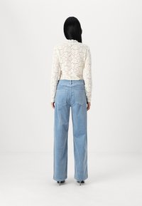 Haut à manches longues en dentelle crème avec un motif floral, associé à un jean en denim bleu clair à jambe large. La vue arrière montre deux poches arrière.