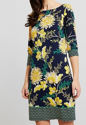Kvinde iført marineblå kjole med gult blomsterprint og grøn mønstret kant på ærmer og kant, stående mod ensfarvet baggrund.