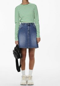 Henkilö, jolla on päällä vihreä-valkoiraidallinen pitkähihaiselta paita, siniset farkkuhameet, valkoiset sukat, valkoiset tennarit, ja joka pitää pientä mustaa reppua sivulla.