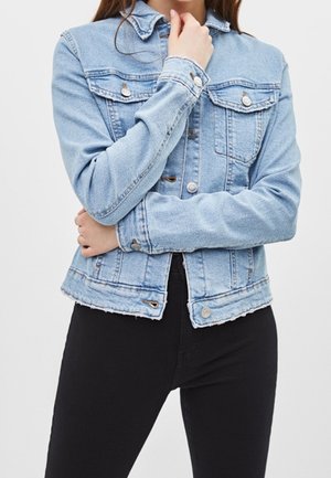 Person wearing a light blue denim jacket with buttoned chest pockets and black pants, arms crossed, hand touching chin.