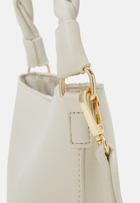 Jasnobeżowa torebka z plecionym uchwytem, złotymi okuciami i teksturowaną skórzaną powierzchnią z widocznymi detalami szwów.