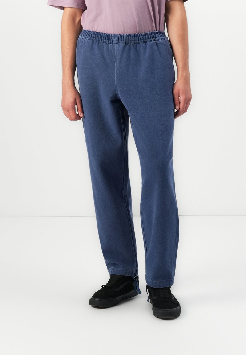 Personen iført løse blå bukser med elastisk midjebånd, en lyse lilla kortermet skjorte, og svarte joggesko som står på et hvitt gulv.
