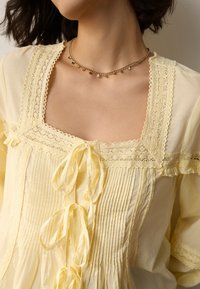 Żółta bawełniana bluzka z pionowymi plisami, koronkowymi zdobieniami na dekolcie i rękawach oraz kokardkami z przodu.