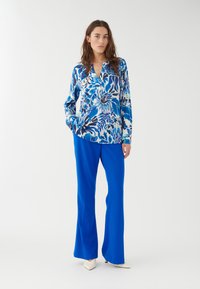 Blusa con motivo floreale in tonalità blu, con maniche lunghe e scollatura a V. Abbinata a pantaloni a campana blu brillante e tacchi bianchi a punta.