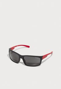Sorte solbriller med en matt finish. Røde stenger har små detaljer. Linsene er mørke og gir UV-beskyttelse. Enkelt, sporty design.