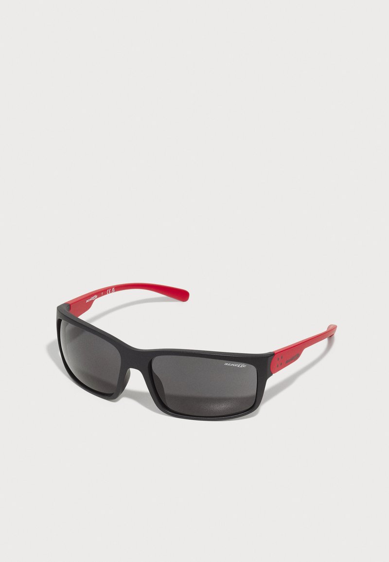 Sorte solbriller med en matt finish. Røde stenger har små detaljer. Linsene er mørke og gir UV-beskyttelse. Enkelt, sporty design.