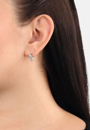 Oreja de mujer con cabello oscuro, que lleva un pequeño pendiente de aro de plata adornado con dos piedras preciosas redondas y transparentes.