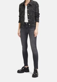 Kobieta w czarno-białej kratkowanej kurtce, białej bluzce, wyblakłych czarnych skinny jeansach i błyszczących czarnych platformach.