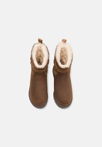 Brune ankelboots med myk hvit pelsfôr, teksturert lærutside, og trekksløyfe bak; gummisåle med grep mønster.
