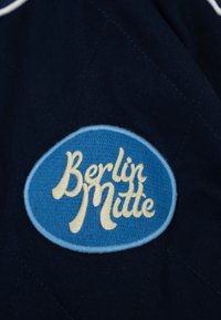 Écusson ovale bleu avec bordure bleu clair et texte crème « Berlin Mitte » cousu sur un tissu bleu marine foncé.