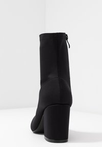 Bottine noire en tissu extensible avec un haut talon bloc. Présente une finition lisse et une fermeture éclair à l'arrière pour un enfilage facile.
