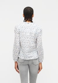 Γυναίκα με πλεξούδες που φοράει λευκό μακρυμάνικο πουκάμισο με μπλε λουλουδάτο σχέδιο και ανοιχτό γκρι εφαρμοστά τζιν, εμφανιζόμενη από πίσω.