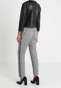 Femme portant un pantalon gris à carreaux avec des bandes latérales noires, une veste courte en cuir noire, et des chaussures à talons hauts bleu marine, debout, dos visible.