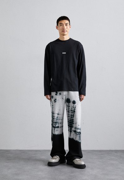 Langärmliges schwarzes Shirt mit zentralem Logo, kombiniert mit grauen und schwarzen Batik-Hosen im weit geschnittenen Stil. Das Model steht auf einem strukturierten grauen Boden.