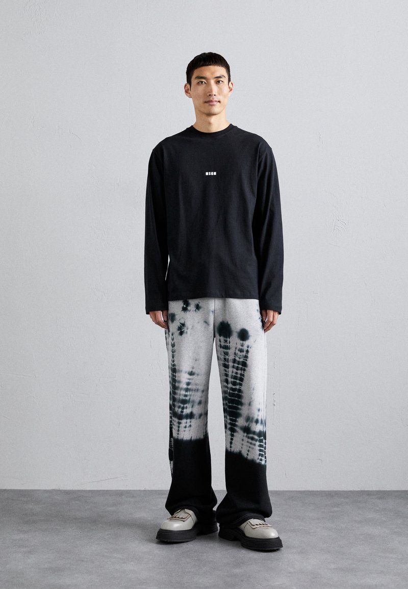 Long-sleeve black shirt with center logo, paired with gray and black tie-dye wide-leg pants. Model stands on textured gray floor.