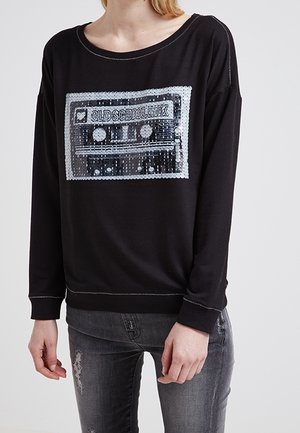 Osoba ubrana w czarną bluzę z naszywką kasety z cekinami z napisem „OLD SCHOOL MIX” oraz zniszczone ciemnoszare dżinsy.