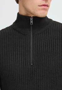 Gebreide zwarte trui met een hoge kraag en een halflange ritssluiting aan de voorkant, voorzien van een gestructureerd patroon en een glanzend ritsdetail.