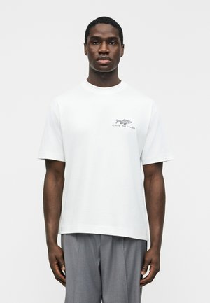 Άνδρας που φοράει λευκό t-shirt με κοντό μανίκι, με γραφικό ψαριού και κείμενο «Κλειστά για το Καλοκαίρι», συνδυασμένο με γκρι παντελόνι, στέκεται μπροστά σε απλό φόντο.