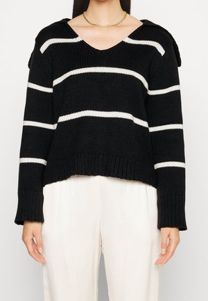 Femme portant un pull en maille noir avec des rayures horizontales blanches et un pantalon blanc, vue des épaules jusqu’aux hanches.