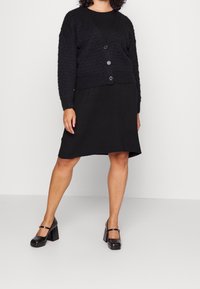 Cardigan noir texturé avec un col en V, trois boutons assortis, porté sur une robe noire simple et associé à des chaussures à talons noires.