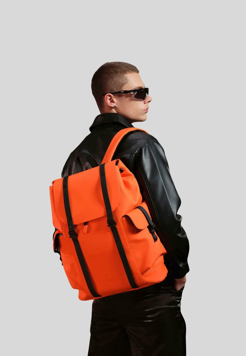 Orange ryggsäck med svarta remmar, strukturerad design, två framfickor och ett handtag på toppen. Bärs tillsammans med en svart läderjacka.