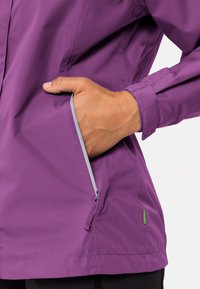 Giacca leggera viola con finitura testurizzata, dotata di una tasca laterale con zip e un accento verde vicino all'orlo.