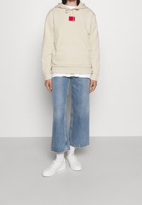 Felpa beige con cappuccio, tasca frontale e patch logo rosso, abbinata a jeans a gamba larga azzurri e sneakers bianche.