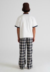 Man wearing a brown beanie, white short-sleeve shirt with black trim, and black and white plaid pants, standing facing away on a plain background.