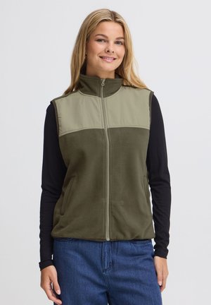 Femme souriante portant un gilet polaire vert zippé sans manches par-dessus une chemise noire à manches longues et un jean bleu sur un fond uni.
