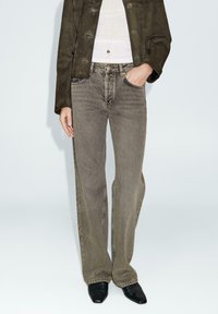 Jeans grigi ampi con texture sbiadita e design a cinque tasche, abbinati a un top bianco e una giacca verde scuro, indossati con scarpe nere a punta.
