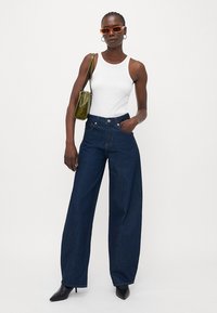 Jeans in denim blu scuro a gamba larga con vita alta, abbinati a una canotta bianca. Gli accessori includono occhiali da sole arancioni e una borsa verde.