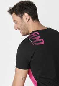Juodas trumpo rankovio marškinėliai su rožinės spalvos grafika nugarinėje pusėje. Marškinėliai turi lenktą apačią ir šonines pink spalvos detales.