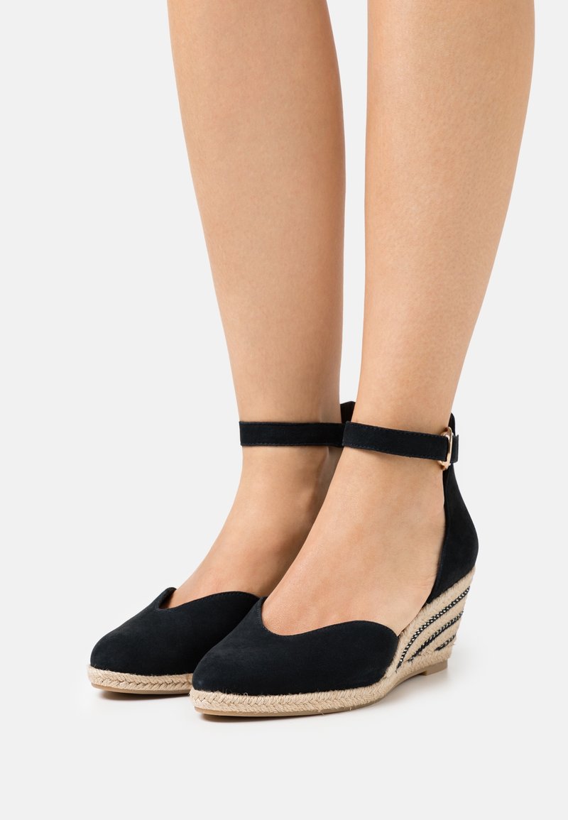 Černé semišové espadrilky na klínu s pásky kolem kotníků, nošené na nohou, s pletenými jutovými podrážkami a dekorativním prošitím na podpatcích.