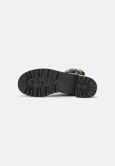 Black footwear with a textured rubber sole featuring a lug design for traction. Accents include gold hardware and adjustable straps.