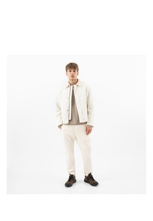 Junger Mann stehend, trägt eine weiße Jacke, einen beigen Pullover, helle Hosen und dunkle Turnschuhe vor einem schlichten weißen Hintergrund.