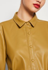 Giacca di pelle color senape con colletto, bottoni a pressione e una texture liscia, caratterizzata da un design aderente e cuciture sottili.
