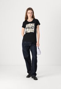 Camiseta negra con un estampado gráfico colorido que presenta un diseño cuadrado. Llevada con jeans de mezclilla oscuros de corte holgado y zapatos negros.