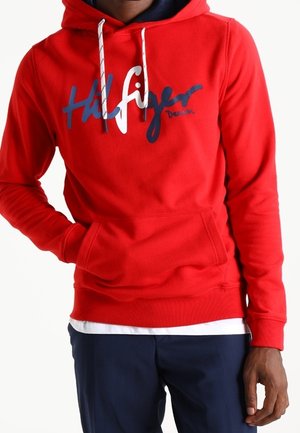 Mężczyzna w czerwonej bluzie z kapturem Tommy Hilfiger z białymi sznurkami i granatowymi spodniami, ręka w kieszeni bluzy.