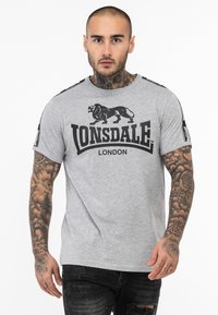 Szürke, rövid ujjú póló fekete oroszlán grafikával és "LONSDALE LONDON" felirattal. Kontrasztos vállcsíkokkal és kerek nyakkivágással rendelkezik.