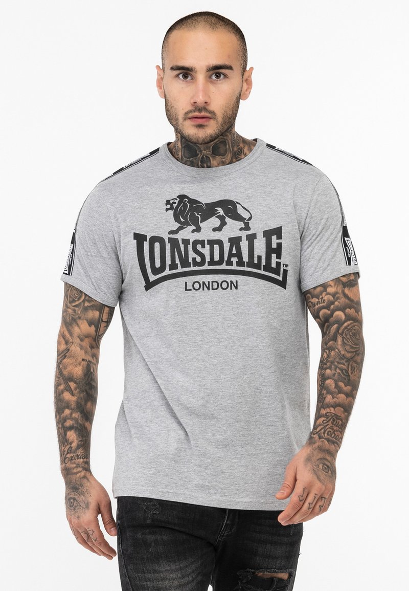 Szürke, rövid ujjú póló fekete oroszlán grafikával és "LONSDALE LONDON" felirattal. Kontrasztos vállcsíkokkal és kerek nyakkivágással rendelkezik.