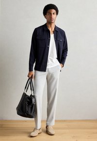 Veste à boutons marine, T-shirt blanc, pantalons gris clair et mocassins beiges. Tenant un sac noir à bandoulière. Fond en parquet en bois.