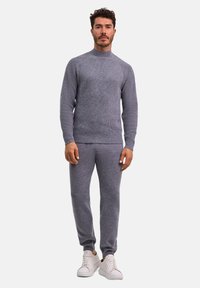 Mann steht und trägt einen grauen Strickpullover und passende Hose mit weißen Turnschuhen vor einem schlichten weißen Hintergrund.
