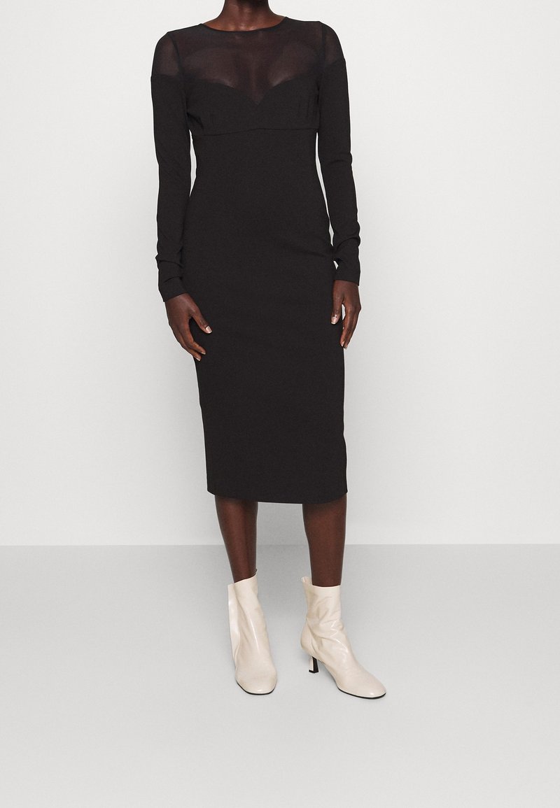 Robe noire à manches longues avec un panneau supérieur transparent, une silhouette ajustée, longueur au genou, complétée par des bottines crème.