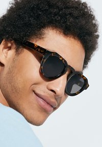 Gafas de sol de carey con lentes redondos, que presentan un grueso marco y acentos laterales. Los lentes son oscuros y ligeramente reflectantes.