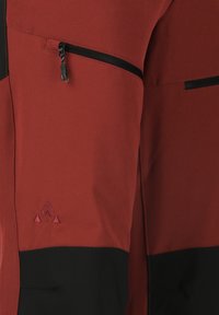 Röda och svarta byxor med en dragkedjeficka, tillverkade av ett texturerat tyg. Har en triangulär logotyp i rött på sidan.