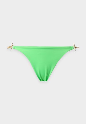Donna in piedi scalza con la mano sul fianco che indossa un bikini verde chiaro con dettagli a catena dorata su sfondo bianco semplice.
