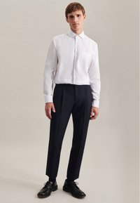 Camicia bianca a maniche lunghe con taschino sul petto, abbinata a pantaloni blu scuro. Scarpe eleganti nere completano l'outfit formale.